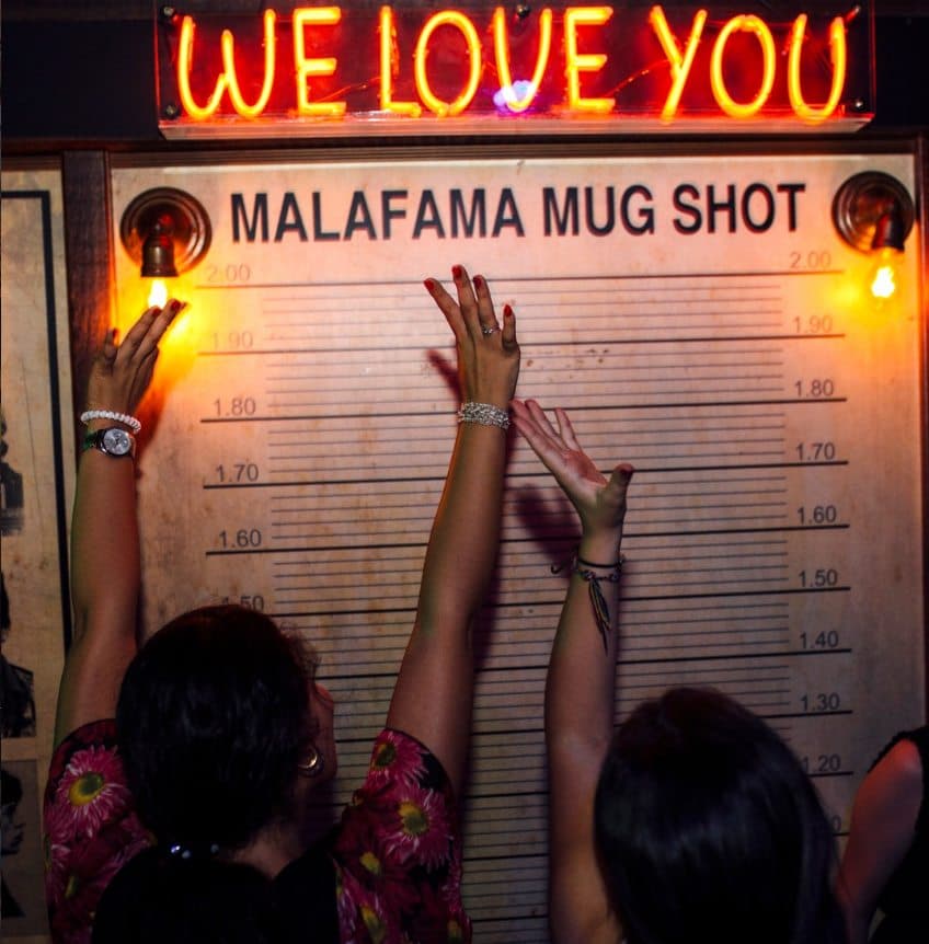 Clientas posando ante la escala de altura del MALAFAMA MUG SHOT bajo el neón WE LOVE YOU